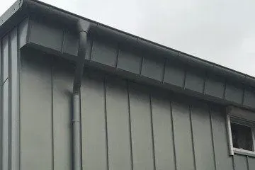 Graue Metalldach- und Wandverkleidung mit einem vertikalen Fallrohr an der Seite eines modernen Gebäudes.