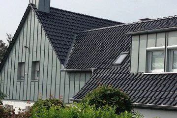 Ein Haus mit dunklem Ziegeldach und grauer, vertikaler Metallverkleidung an Giebel und Dachgaube.