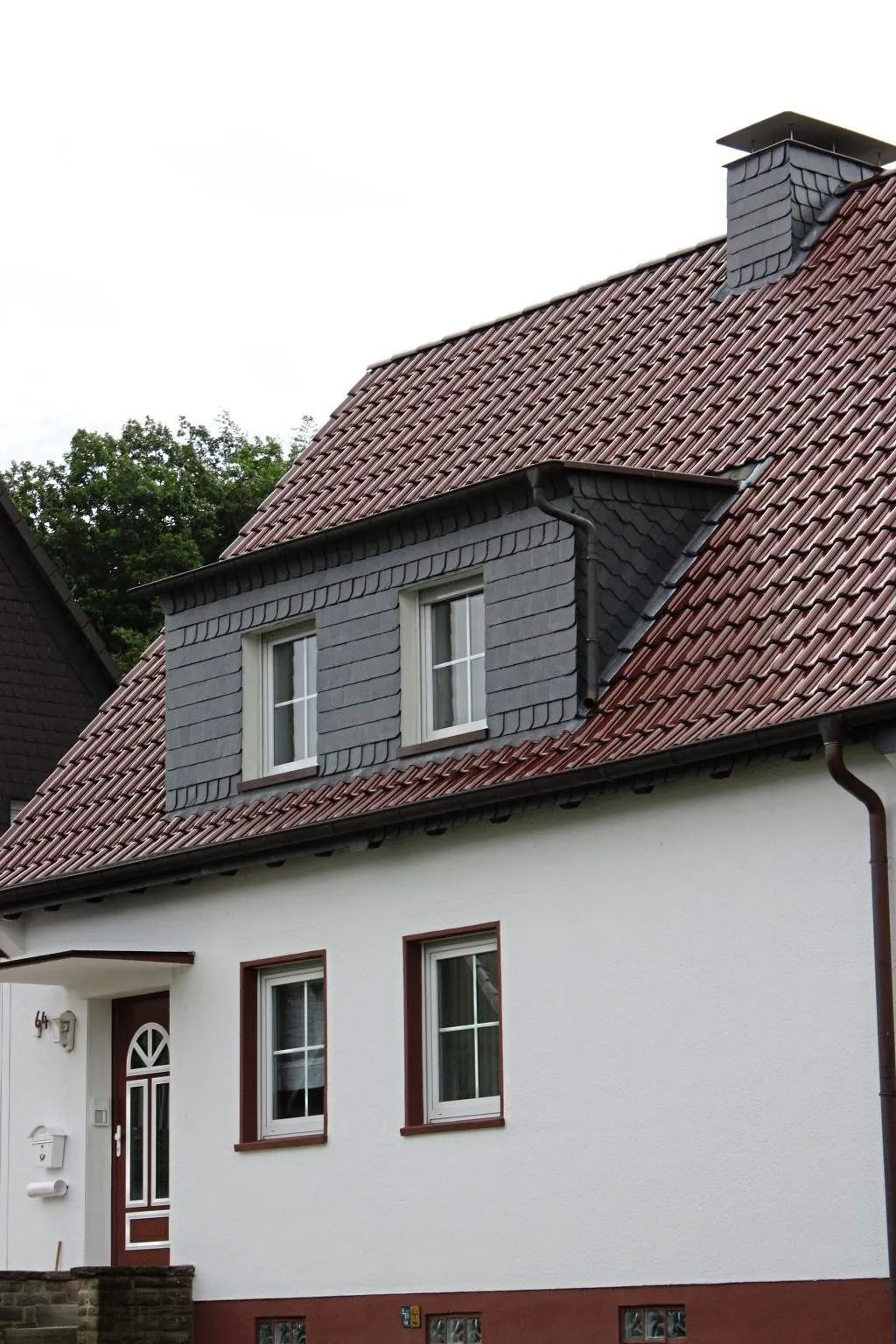 Weißes Haus mit rotem Ziegeldach, einem grauen Dachgaubenfenster mit zwei Fenstern und einer Eingangstür.