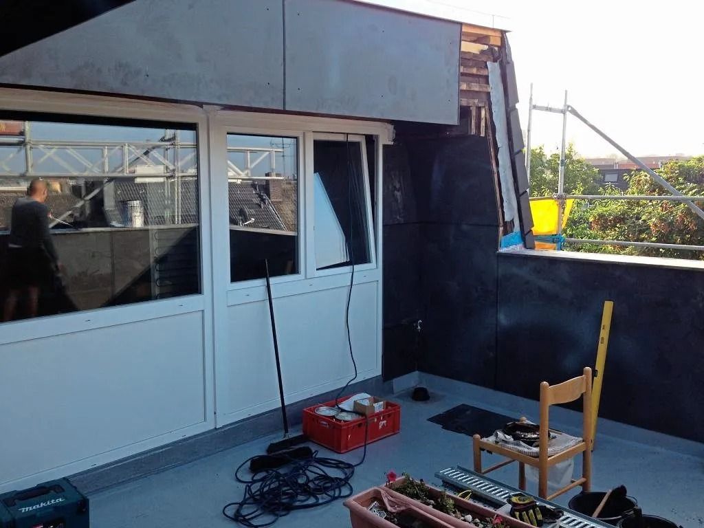 Eine im Bau befindliche Dachterrasse mit weißen Fenstern, Werkzeugen und einem Holzstuhl vor einer dunklen Wand.