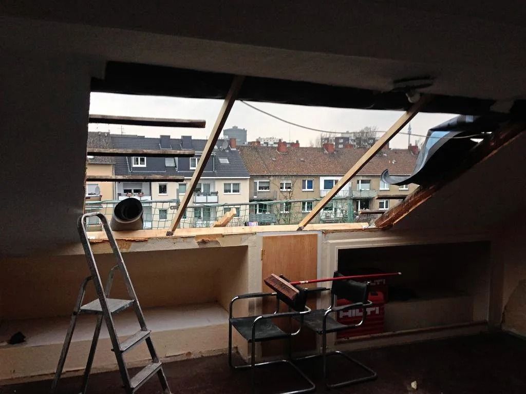 Ein im Bau befindliches Dachzimmer mit offenem Dach, einer Leiter und zwei Stühlen mit Blick auf die Stadtgebäude.