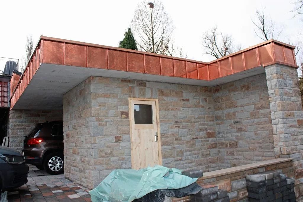 Steingarage mit Holztür und rostfarbenem Dachrand, darin geparktes Auto und davor Baumaterialien