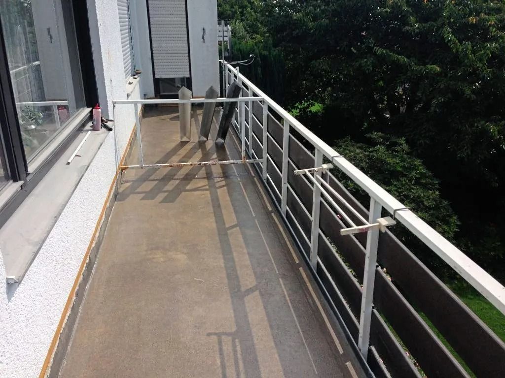 Ein langer, schmaler Außenbalkon mit weißem Geländer, einem braunen Sichtschutz aus Holz und einigen Wäscheständern.