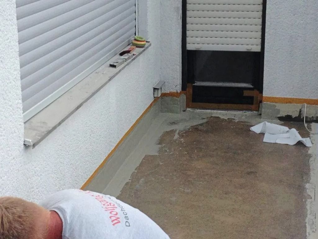 Ein Arbeiter trägt wasserdichtes Dichtmittel auf eine Ecke eines Außenbalkons in der Nähe einer Tür und eines Fensters auf.