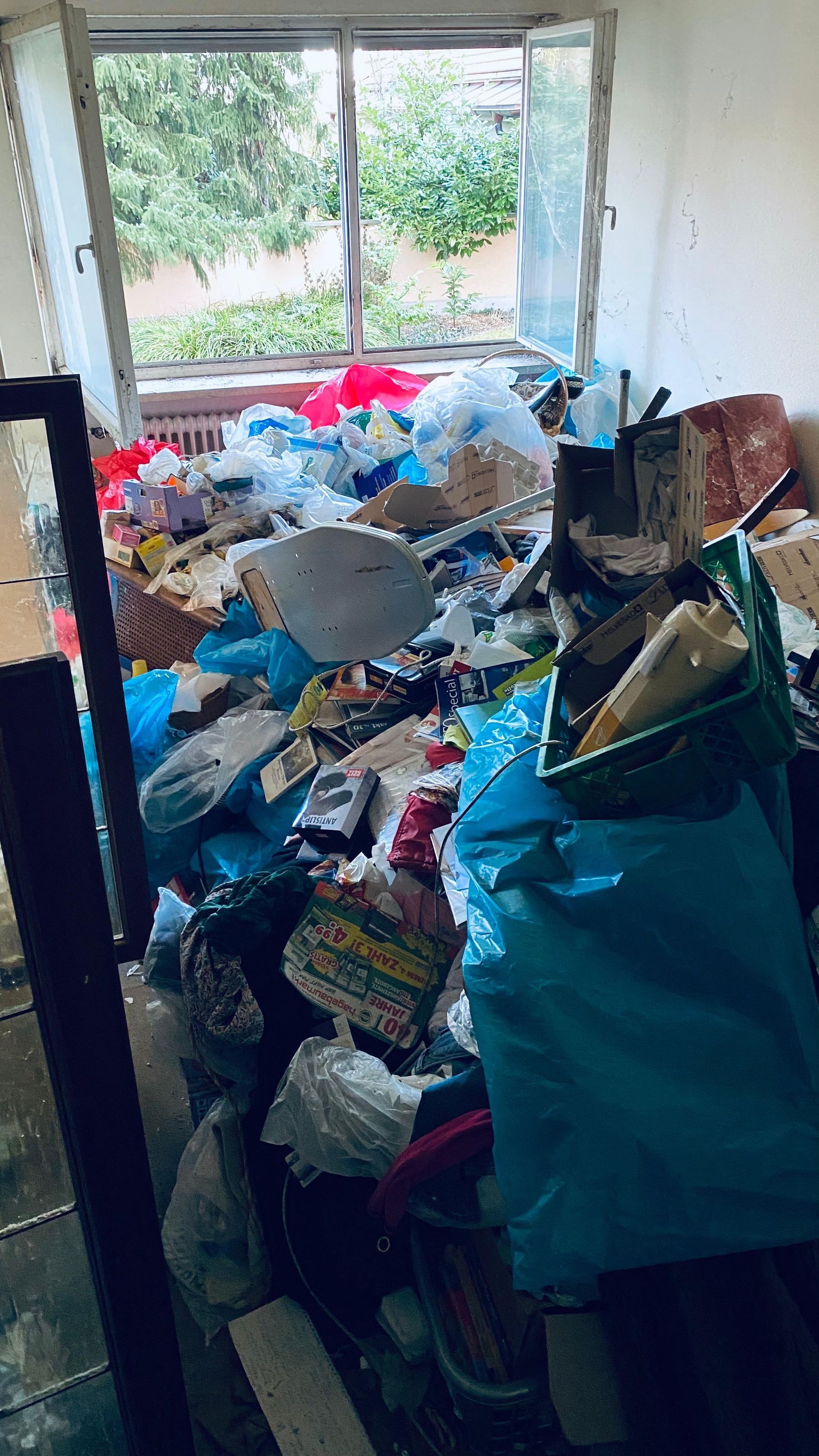 Unaufgeräumtes Zimmer mit Blick aus dem Fenster auf Bäume.