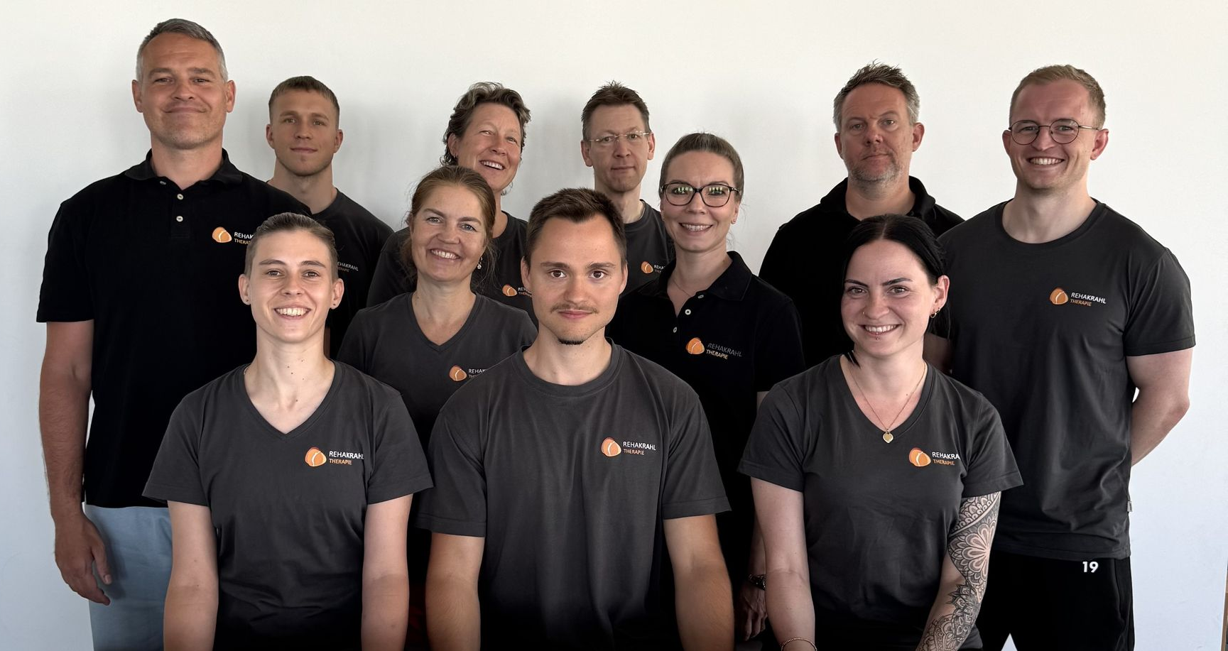 Eine Gruppe von 12 Personen in dunklen Hemden posiert vor einer weißen Wand.