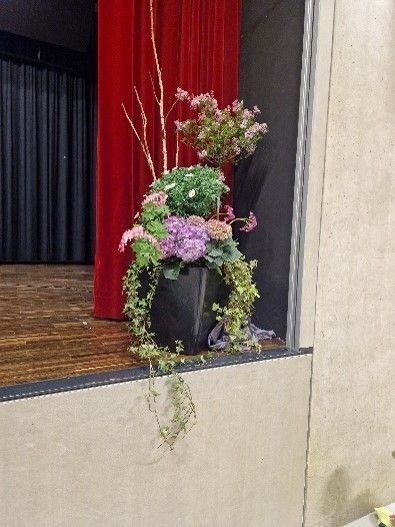 Auf einer Bühne vor einem roten Vorhang steht eine mit Blumen gefüllte Vase.