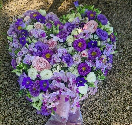 Auf dem Boden liegt ein herzförmiges Arrangement aus violetten und rosa Blumen.