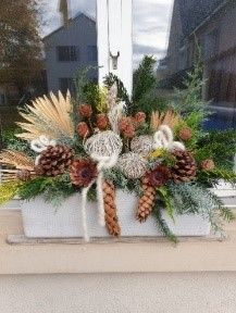 Ein mit Blumen und Tannenzapfen gefüllter Blumenkasten auf einem Fensterbrett.