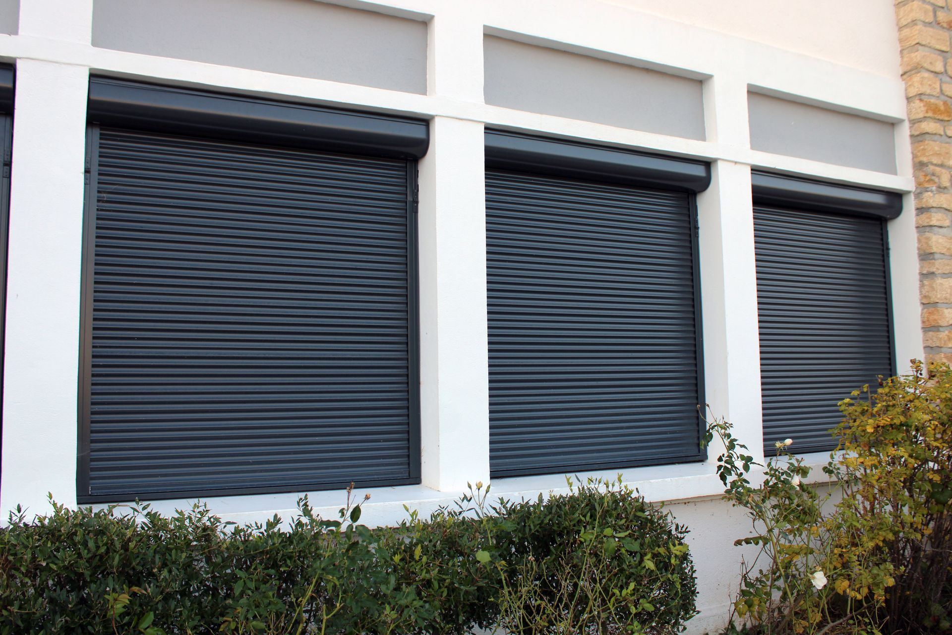 Trois volets roulants de sécurité horizontaux gris foncé recouvrent les fenêtres d'un bâtiment blanc, avec des arbustes en contrebas.