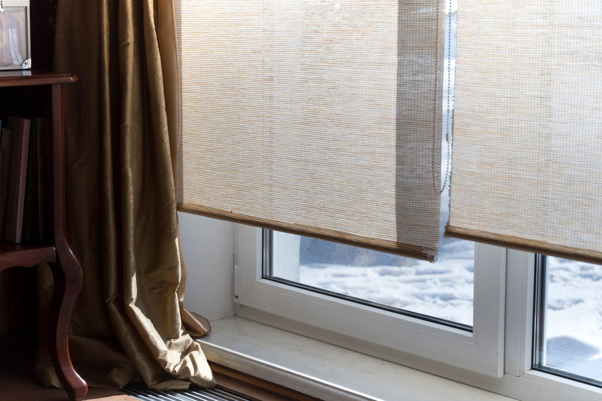 Une fenêtre aux stores texturés brun clair partiellement baissés, à côté d'un rideau doré et d'une étagère en bois foncé.