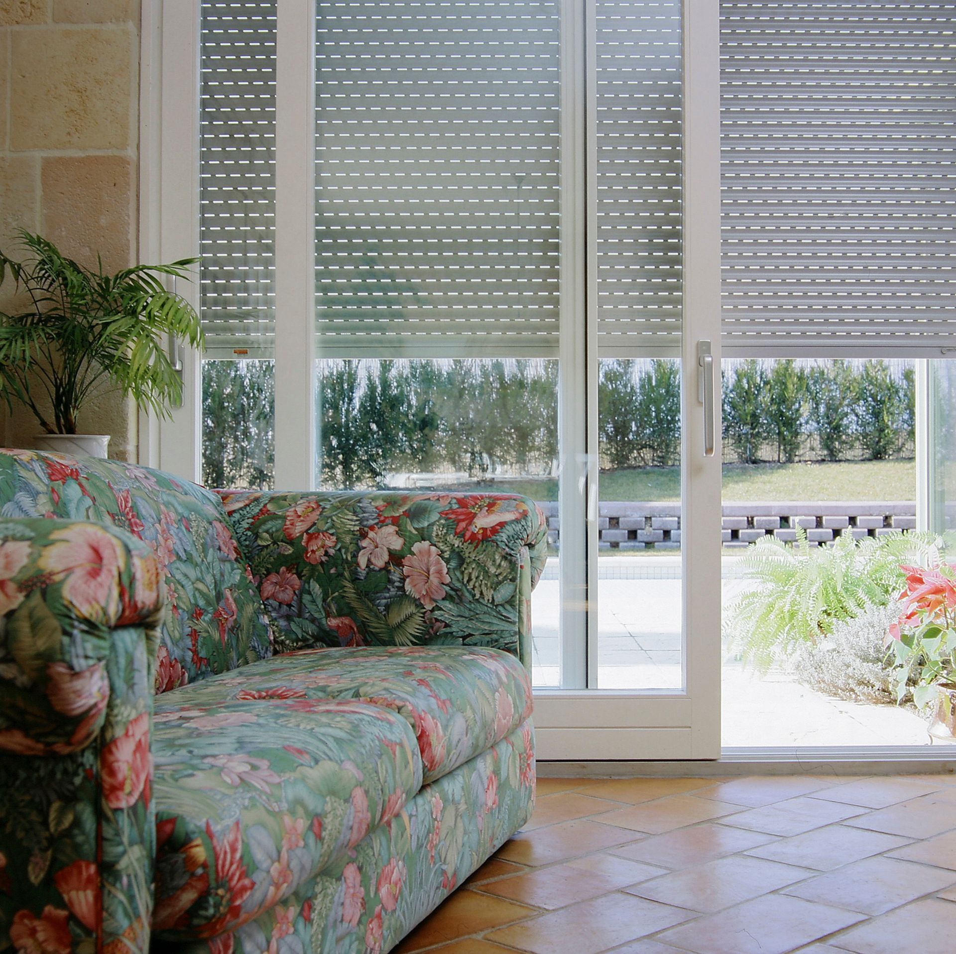 Un canapé à imprimé floral est placé à côté d'une porte vitrée avec stores enrouleurs horizontaux, donnant sur un jardin.