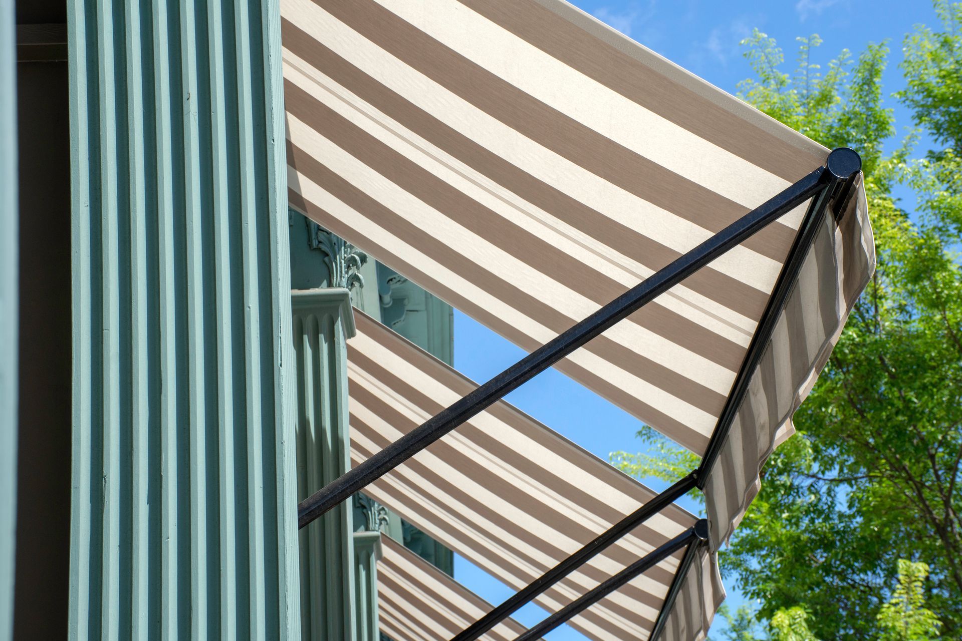 Un store rétractable à rayures beiges et blanches, fixé à un mur nervuré bleu sarcelle, avec des arbres verts sur fond de ciel bleu.