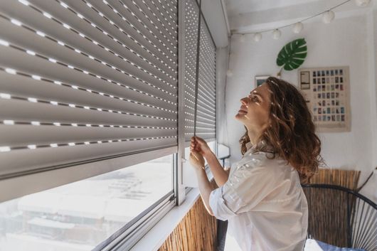 Une personne en chemise blanche se tient près d'une fenêtre, ajustant de ses mains des volets roulants clairs.