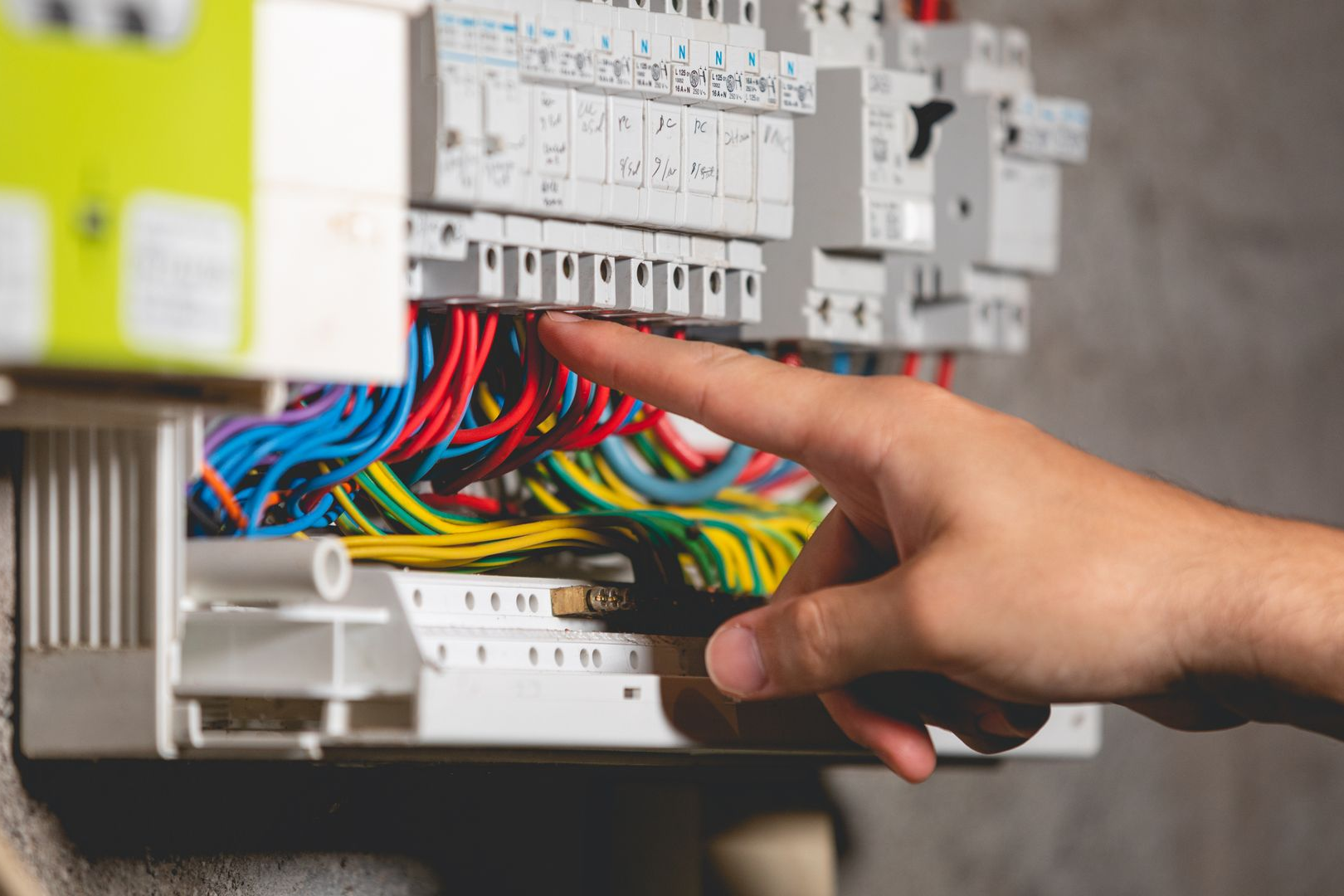 Main pointant des fils électriques à l'intérieur d'un boîtier de disjoncteurs. Des fils rouges, bleus, jaunes et verts sont visibles.