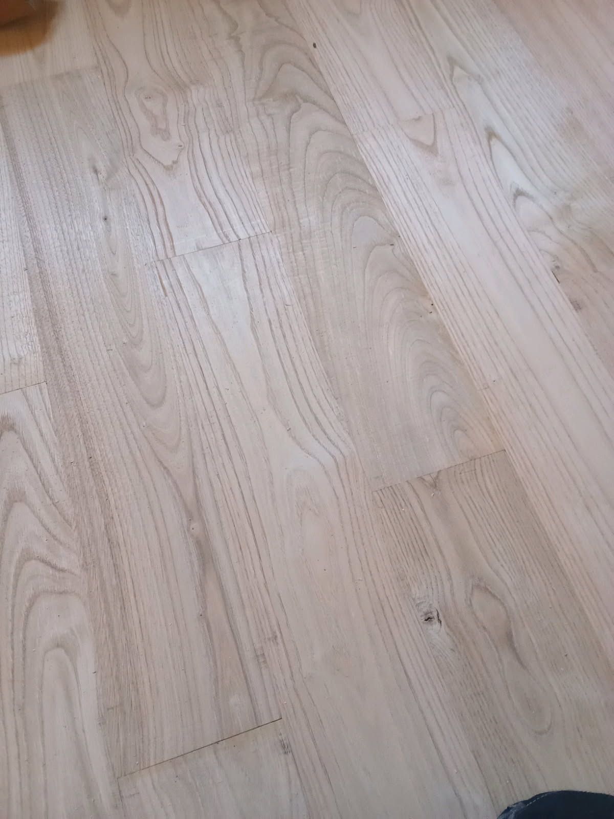 Une personne teint un parquet à chevrons au pinceau ; la moitié du parquet est teintée.
