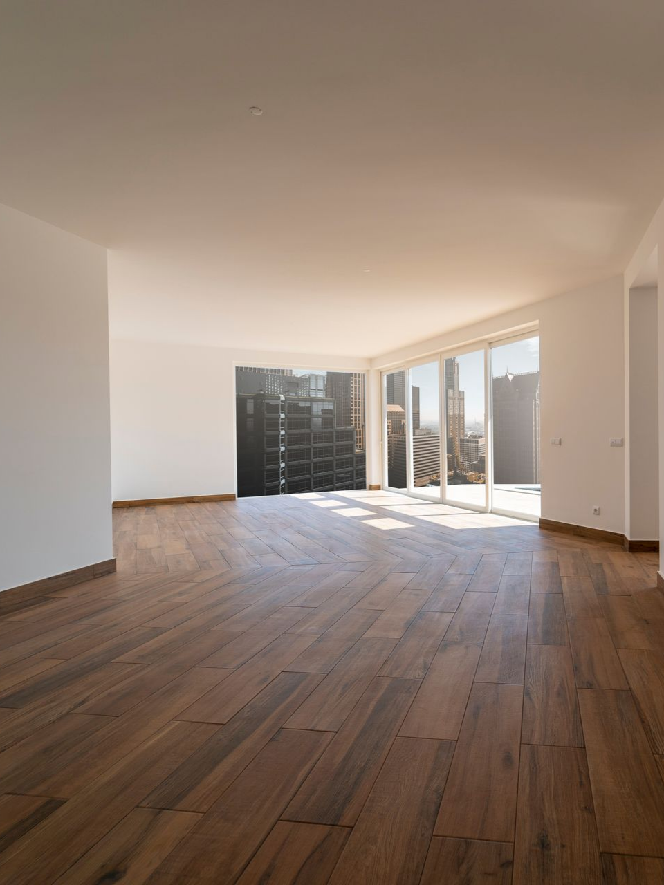 Pièce vide avec parquet, murs blancs et grandes fenêtres offrant une vue sur la ville.
