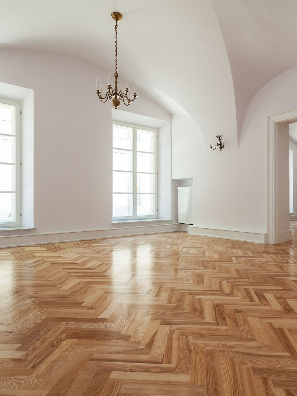 Pièce vide avec parquet clair, murs blancs, plafond voûté et lustre.
