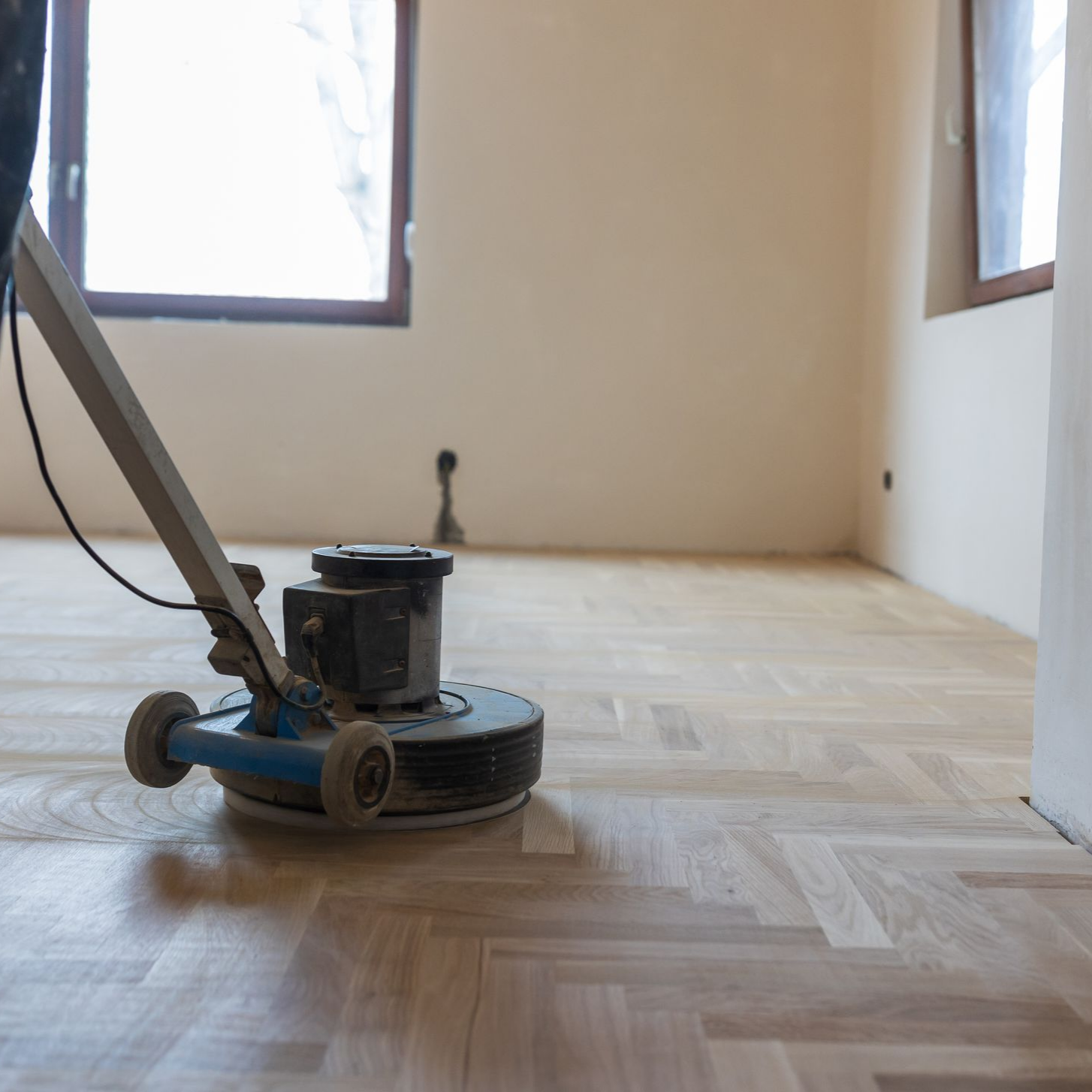 Une ponceuse à parquet polit un plancher de bois franc dans une pièce avec des fenêtres et des murs de couleur claire.