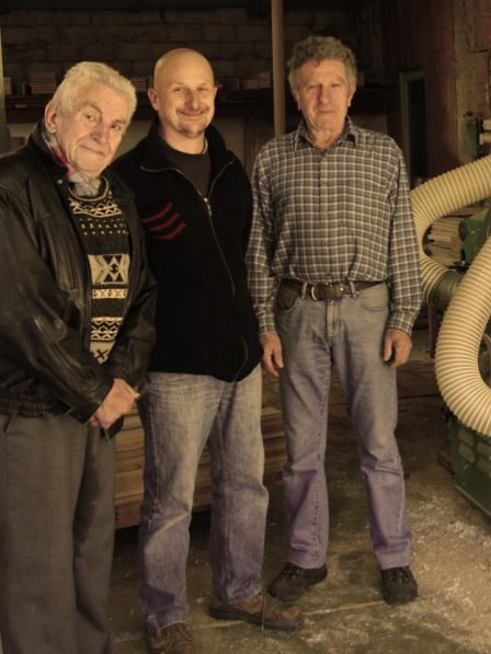 Trois hommes se tiennent dans un atelier. Celui de gauche porte une veste. Les autres portent des pulls et des jeans.