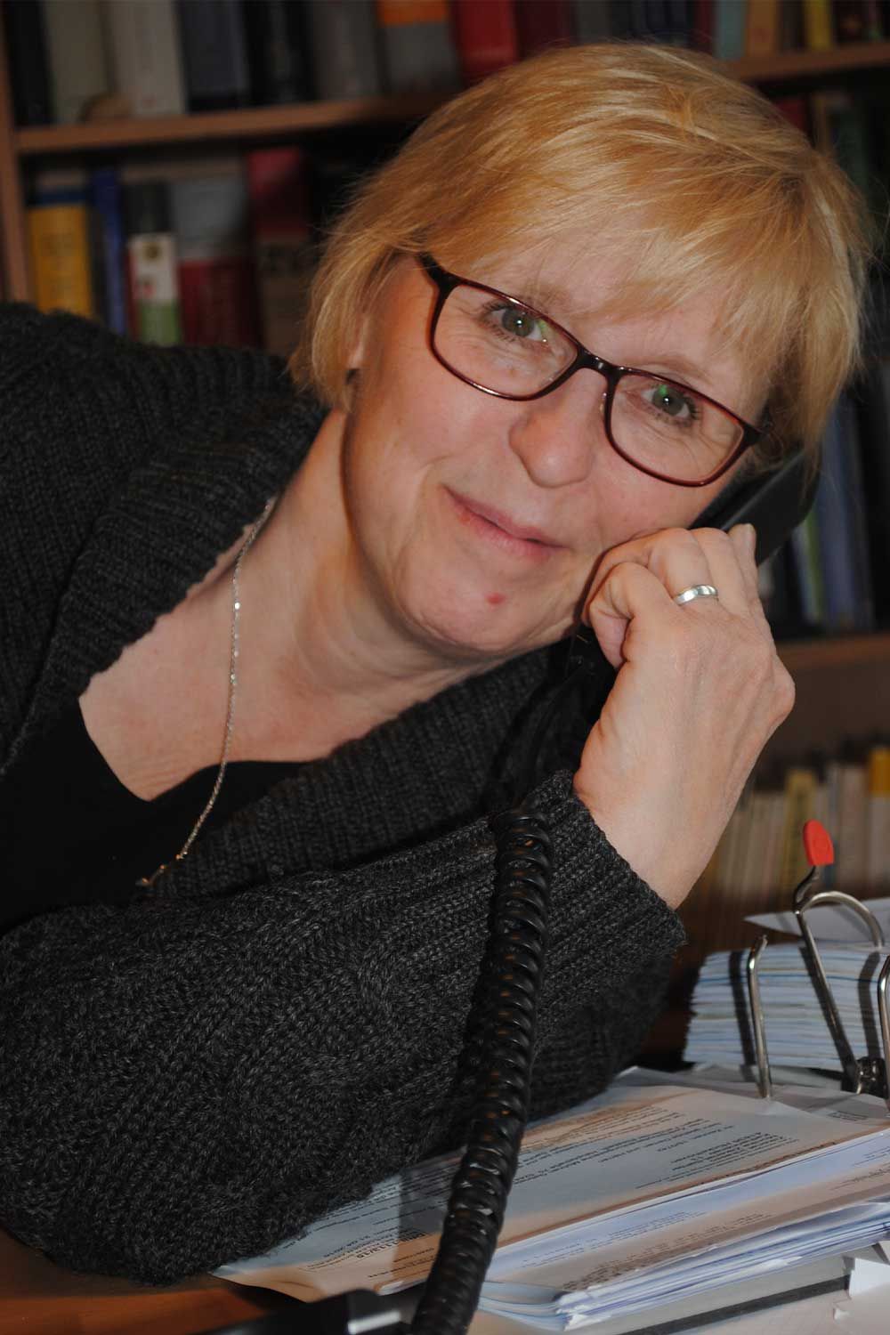 Eine Frau mit Brille sitzt an einem Schreibtisch und hat die Hand am Kinn.
