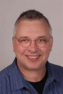 Ein Mann mit Brille und blauem Hemd lächelt in die Kamera.