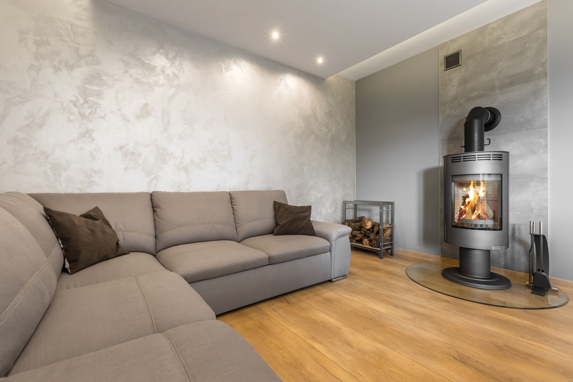 Salon moderne avec un canapé d'angle gris, un parquet et un poêle à bois noir allumé.