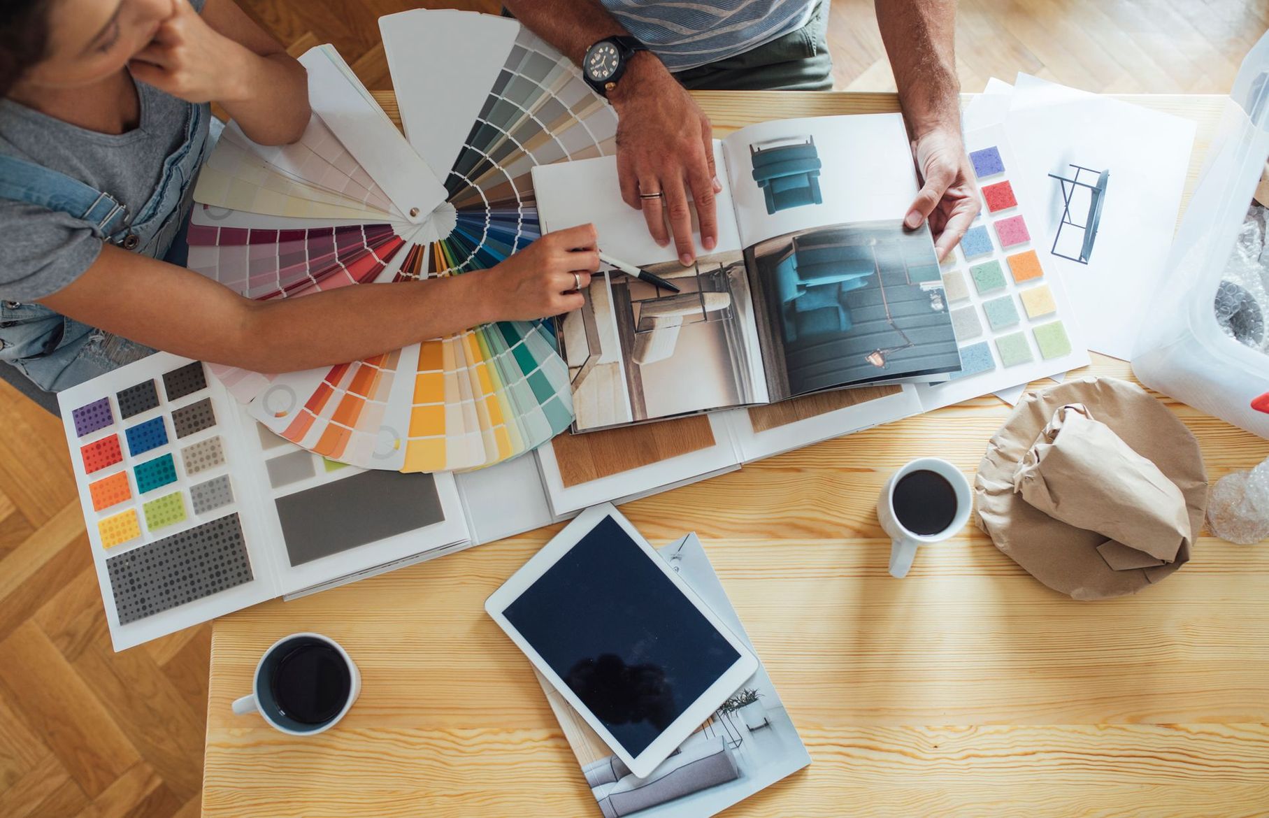 Deux personnes examinent des échantillons de couleurs et une maquette étalée sur une table en bois, avec des tasses à café et une tablette.