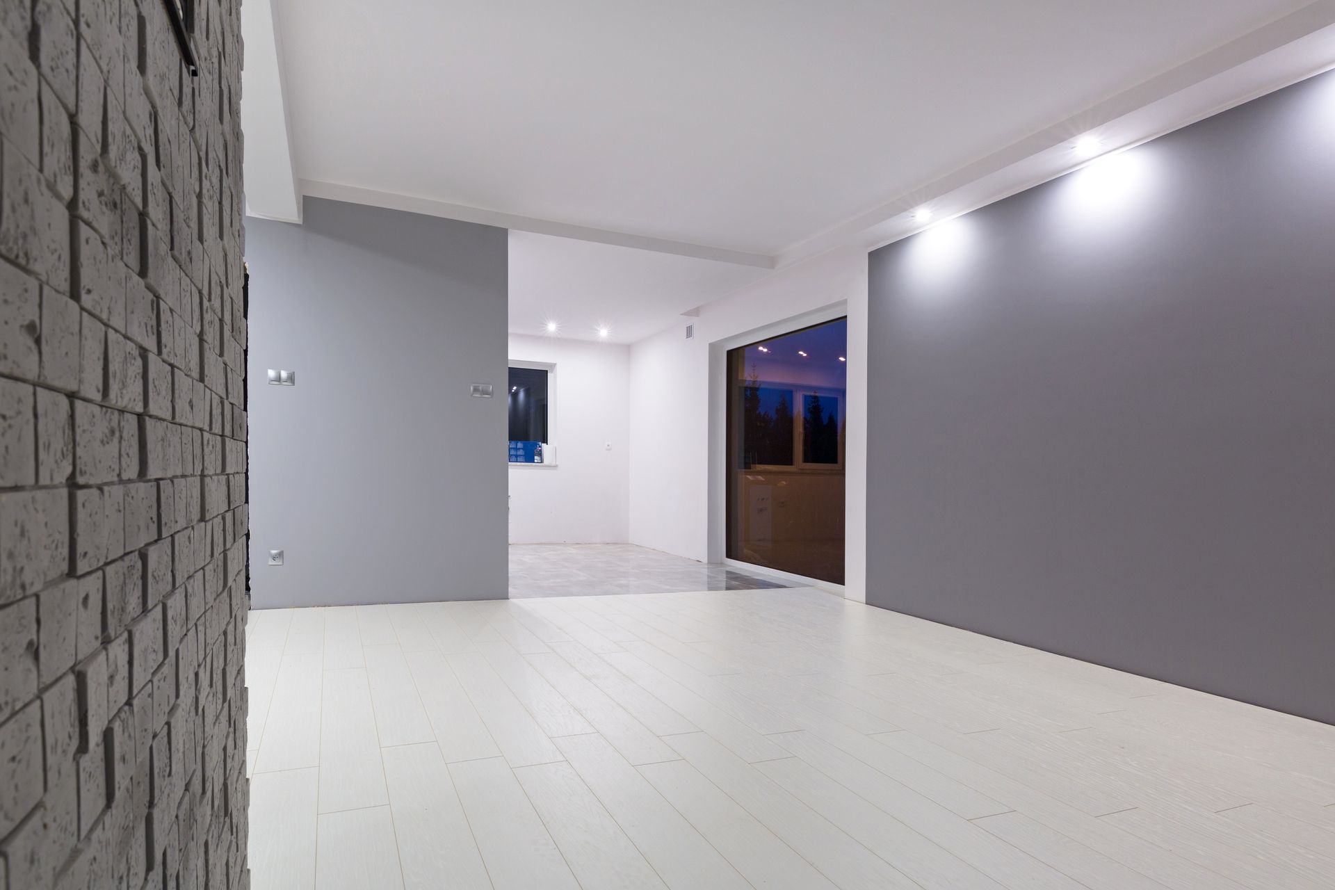 Intérieur moderne et épuré avec sol blanc, murs gris, accents de pierre et porte ouverte sur le crépuscule extérieur