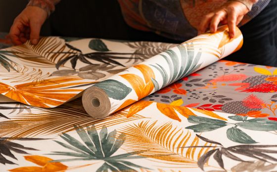 Des mains déroulent un rouleau de papier peint à motifs de feuilles tropicales sur une table