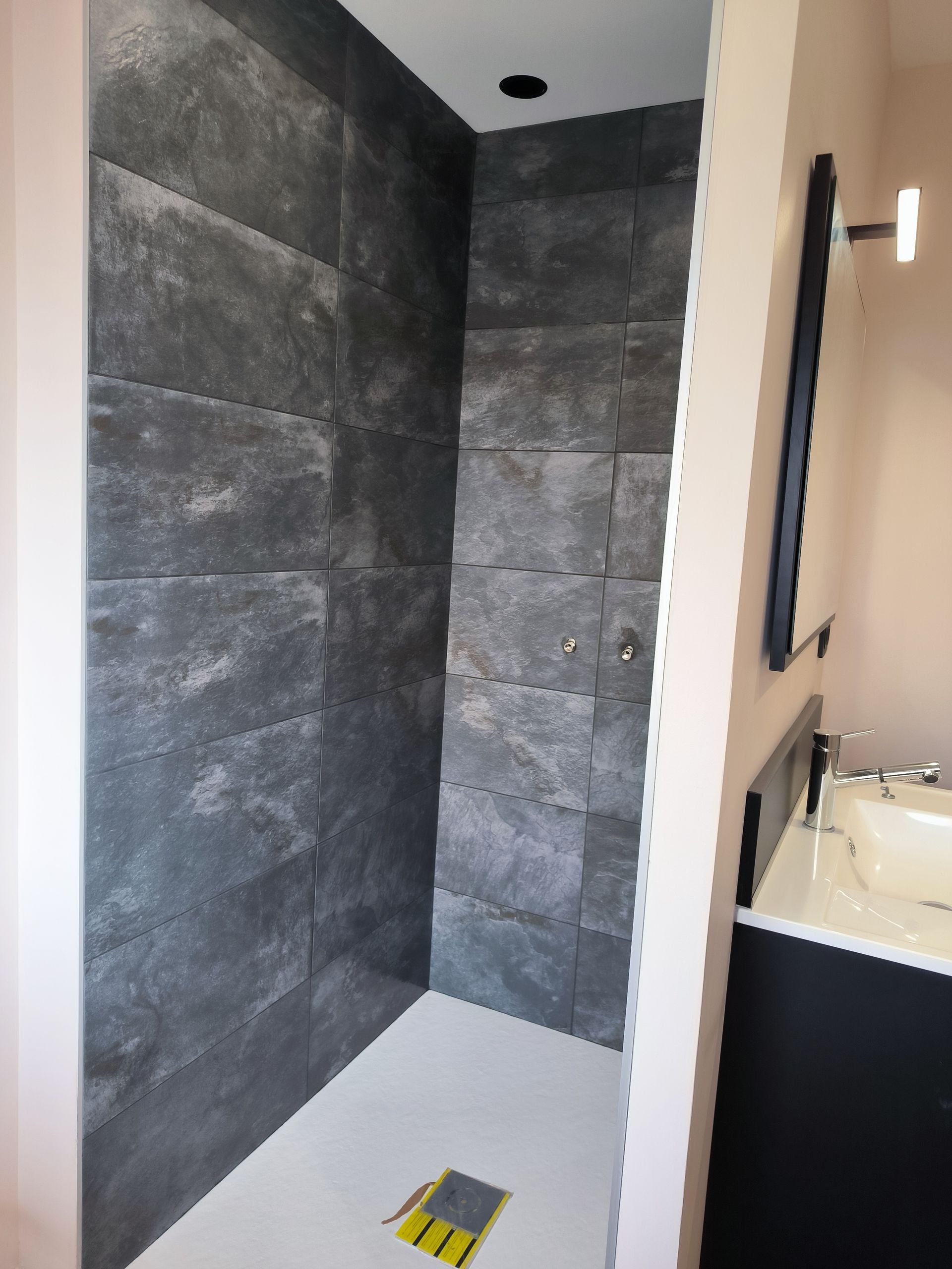 Douche à l'italienne moderne avec murs carrelés de pierre foncée et sol carrelé blanc à côté d'un lavabo.