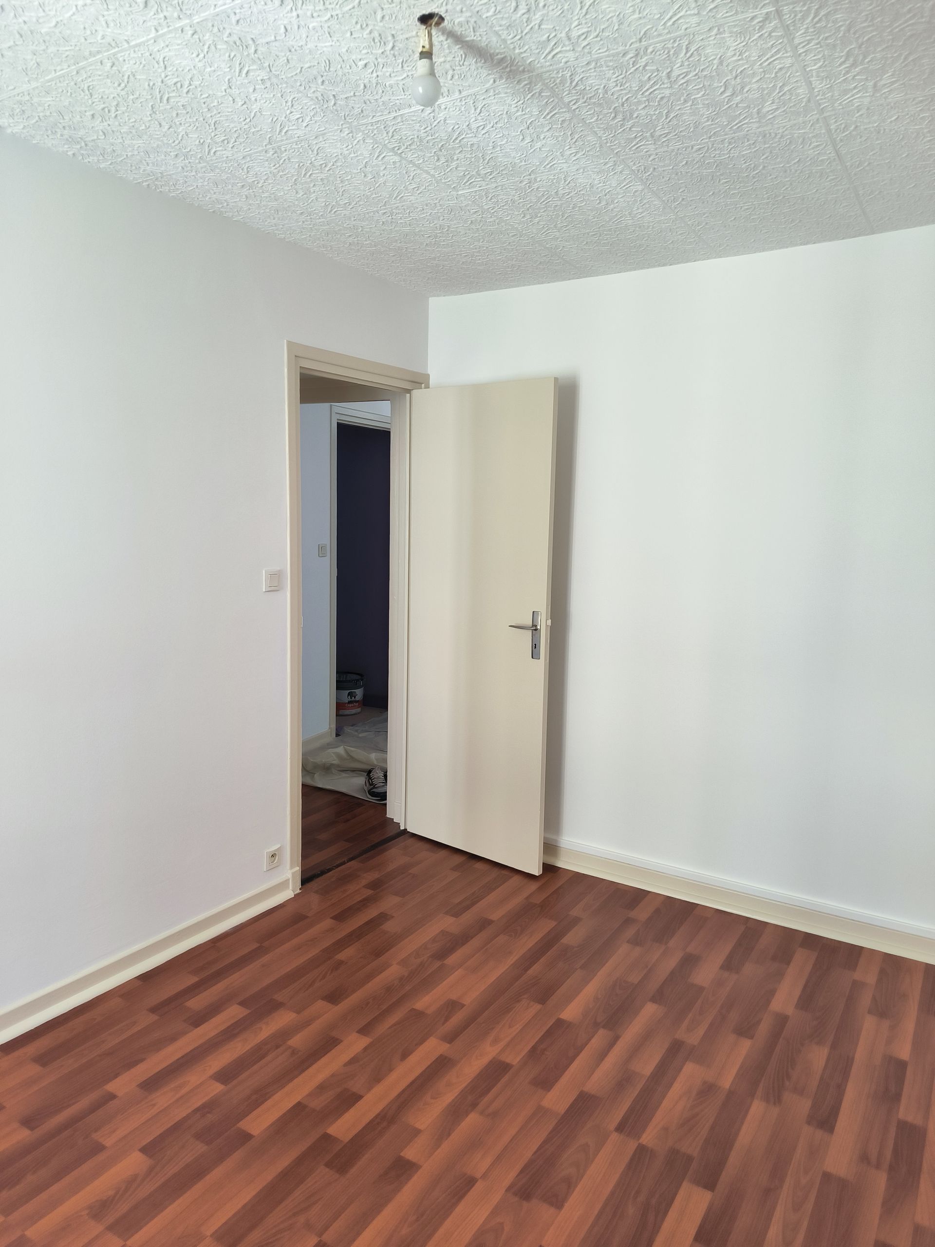 Pièce vide aux murs blancs, plafond texturé, parquet et porte ouverte sur un couloir.