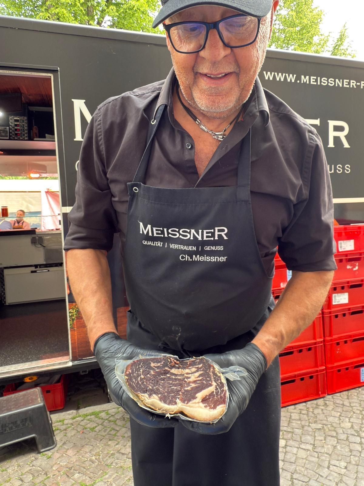 Ein Mann in schwarzer Schürze hält lächelnd ein großes Steak. Hinter ihm steht ein Imbisswagen mit roten Behältern.