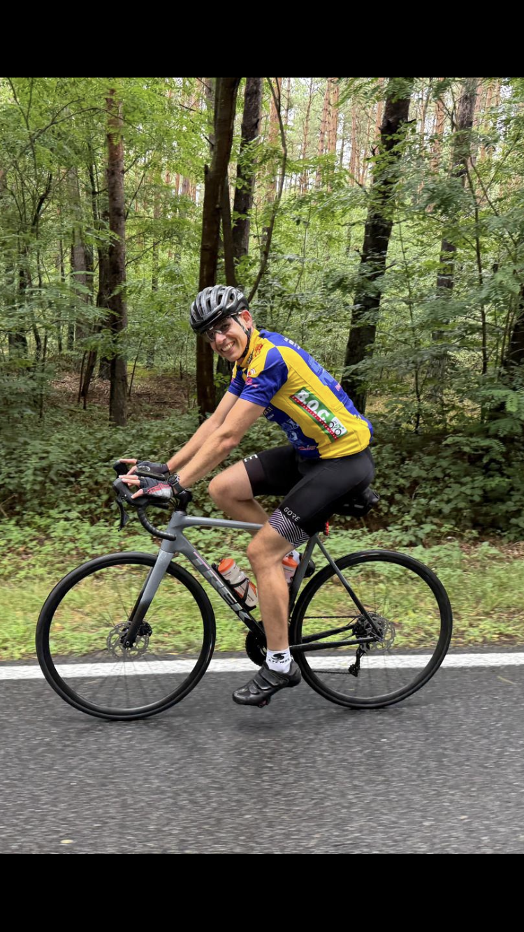 Ein Radfahrer auf einem Rennrad lächelt, während er auf einem asphaltierten Weg durch einen Wald fährt. Er trägt ein blau-gelbes Trikot, einen Helm und schwarze Shorts.