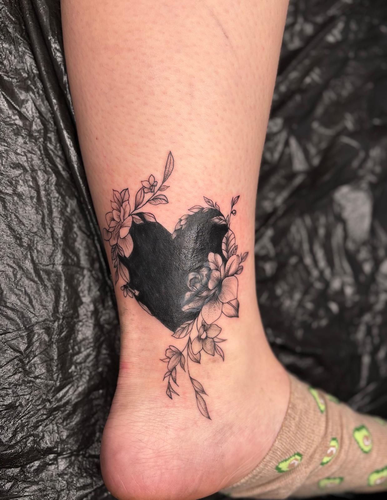 Eine Frau hat ein schwarzes Herztattoo auf ihrem Knöchel, umgeben von Blumen.