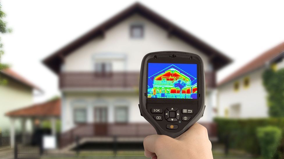 A person is holding a thermal camera in front of a house.
