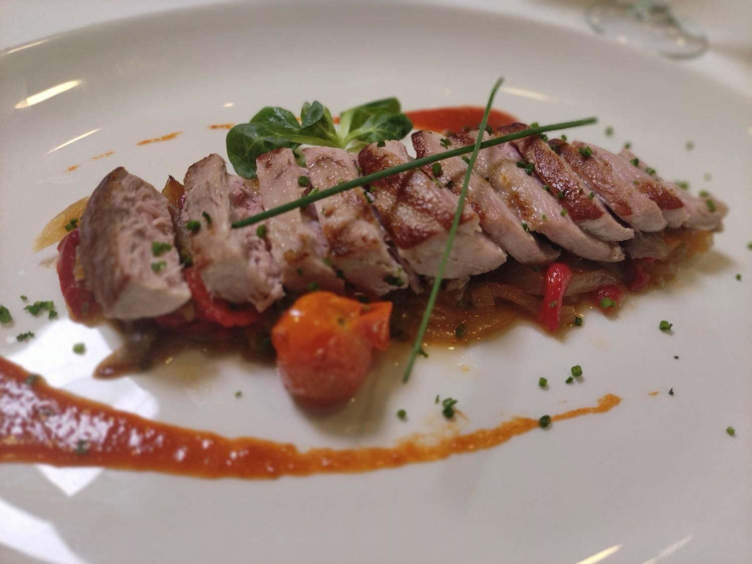 Carne en rodajas sobre verduras asadas, adornada con hierbas y salsa de tomate en un plato blanco.