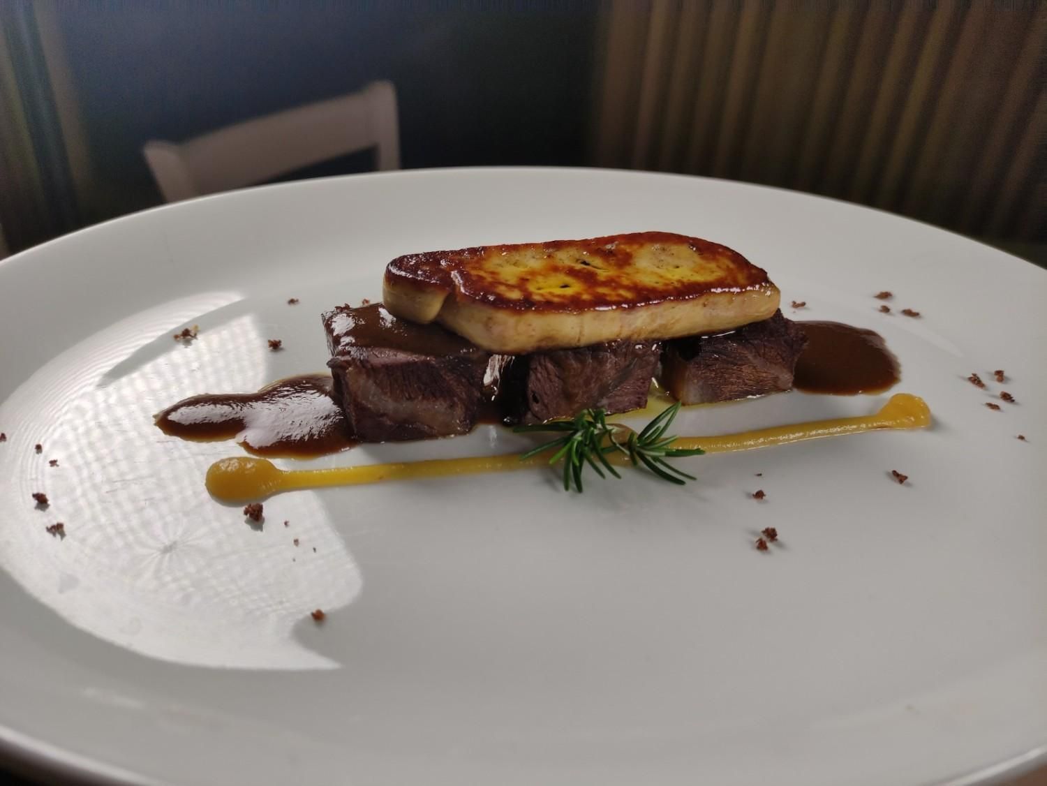 Plato de carne: foie gras sellado sobre cubos de carne apilados, un chorrito de salsa y una ramita de hierbas en un plato blanco.