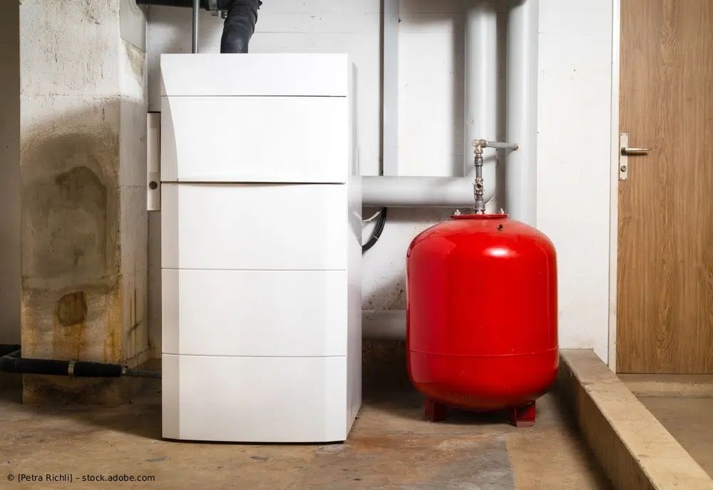 In einem Kellerabteil steht neben einem leuchtend roten Ausdehnungsgefäß ein weißes Heizgerät.