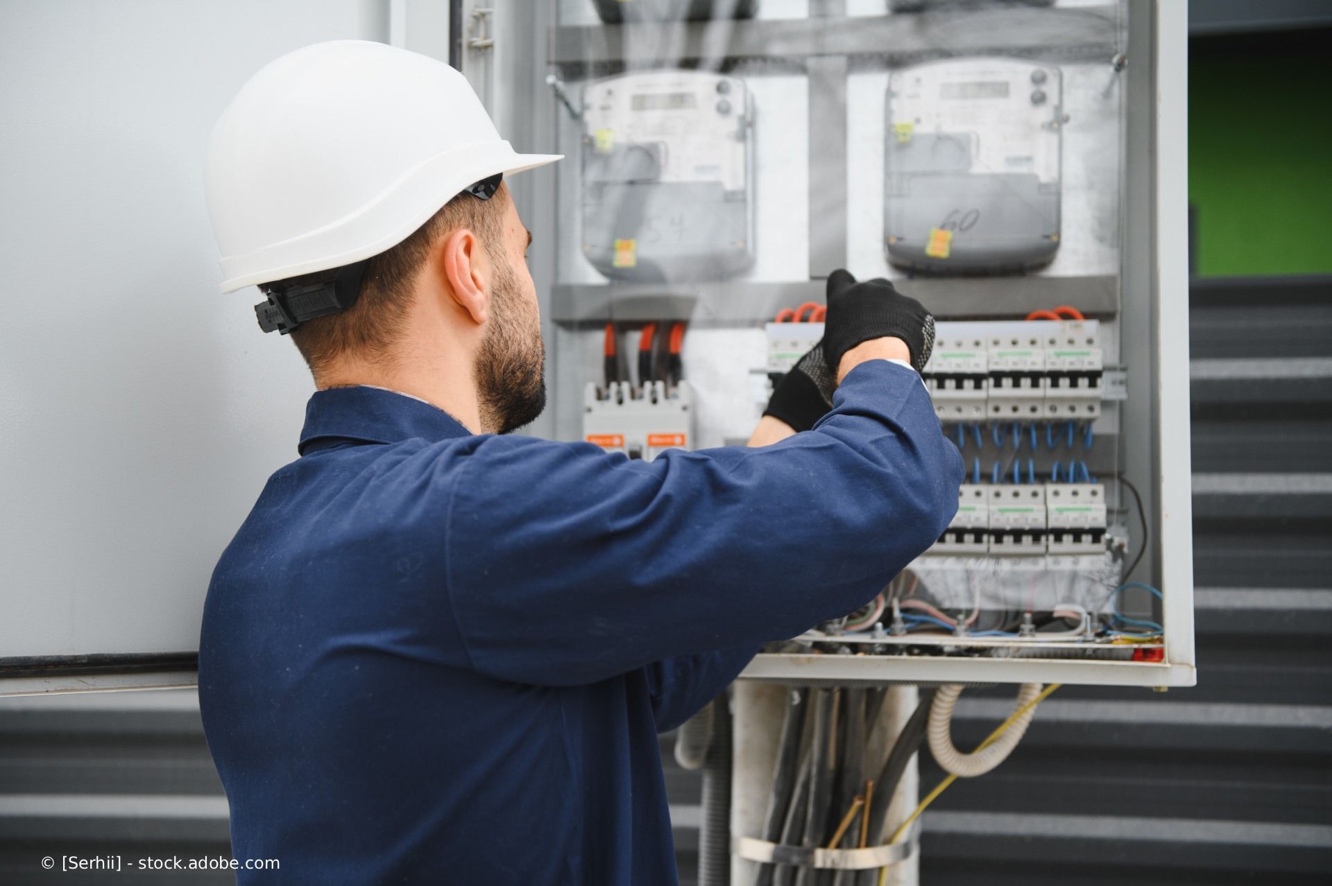 Eine Person, die Arbeitshandschuhe trägt, benutzt einen Schraubendreher mit rotem Griff, um eine an der Wand montierte elektrische Anschlussdose zu installieren oder zu reparieren.