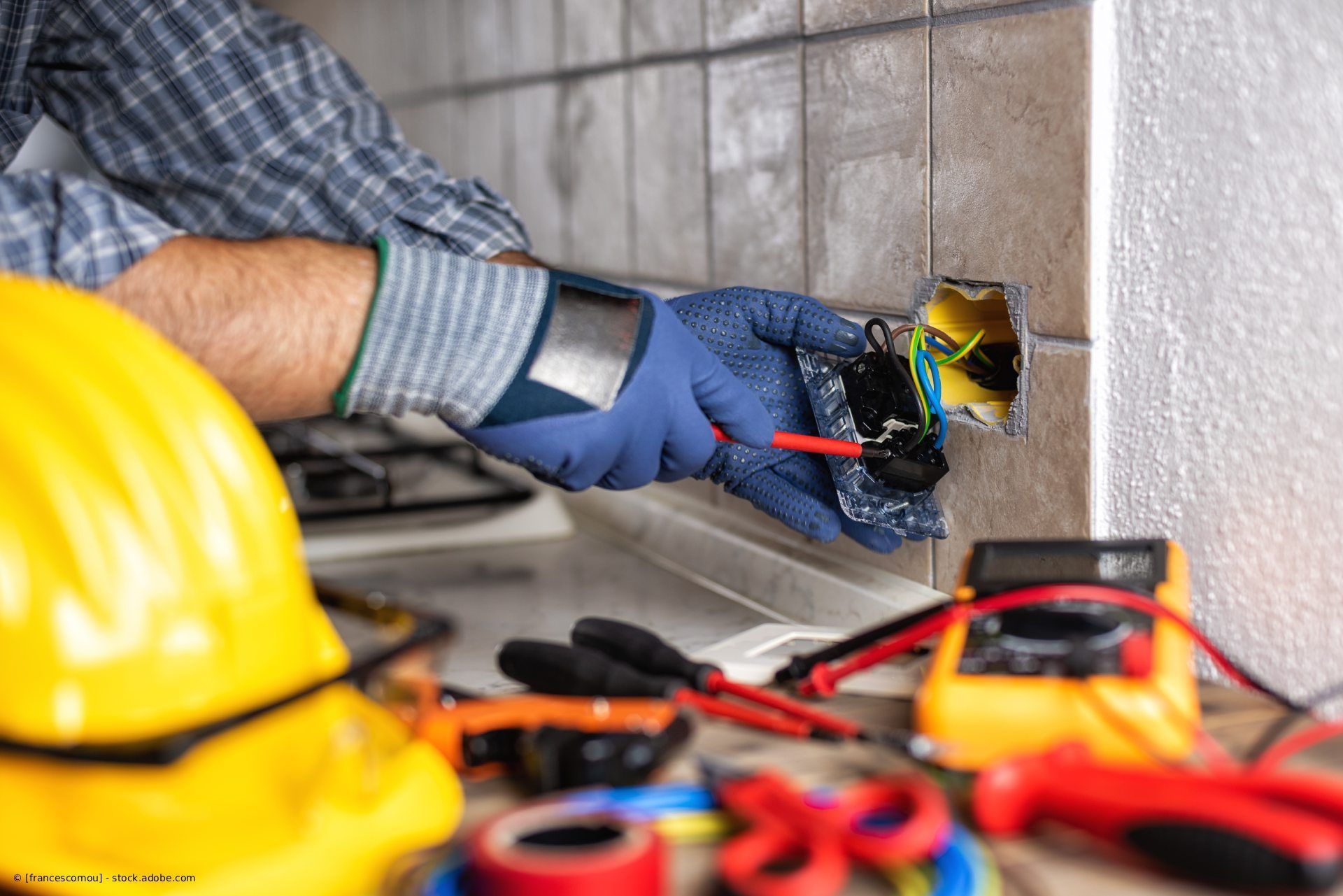 Hände in weißen Arbeitshandschuhen hantieren mit grauen elektrischen Leitungen in der Nähe eines offenen Sicherungskastens an einer Backsteinmauer.