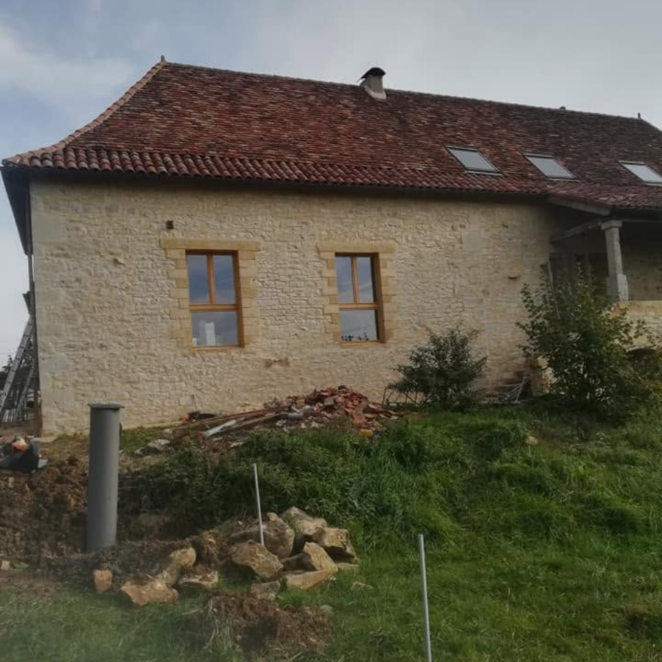 Maison en pierre au toit de tuiles rouges et aux fenêtres en bois, située à flanc d'une colline herbeuse.
