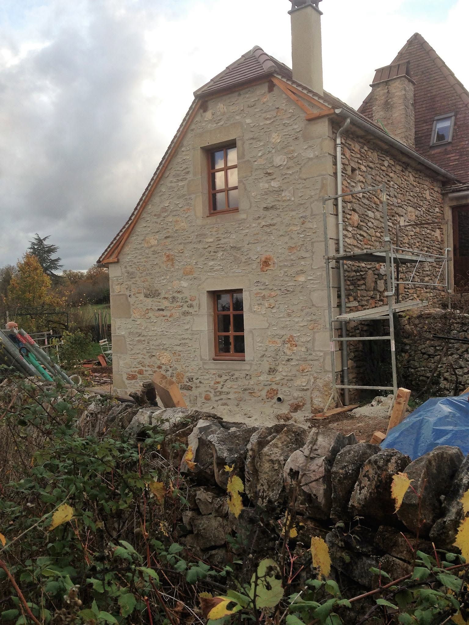 Maison en pierre aux fenêtres encadrées de bois, cheminée et mur de jardin sous un ciel nuageux.