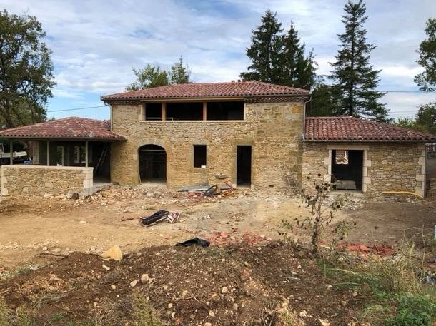 Maison en pierre en construction, avec toit de tuiles rouges, ouvertures cintrées et premier plan en terre battue.