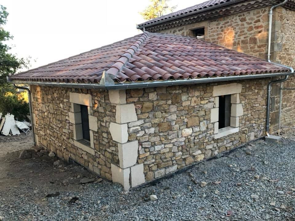 Petite dépendance en pierre avec un toit de tuiles rouges sur un terrain gravillonné, à côté d'un bâtiment en pierre plus grand.
