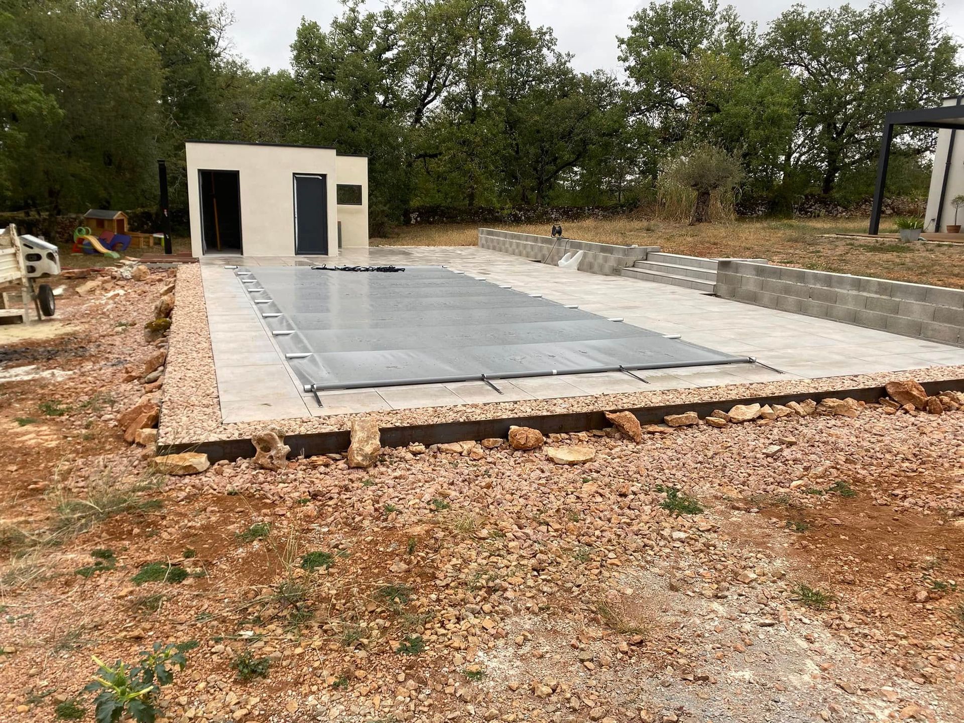 Piscine en béton partiellement construite à côté d'un petit cabanon blanc, entourée de terre et d'arbres