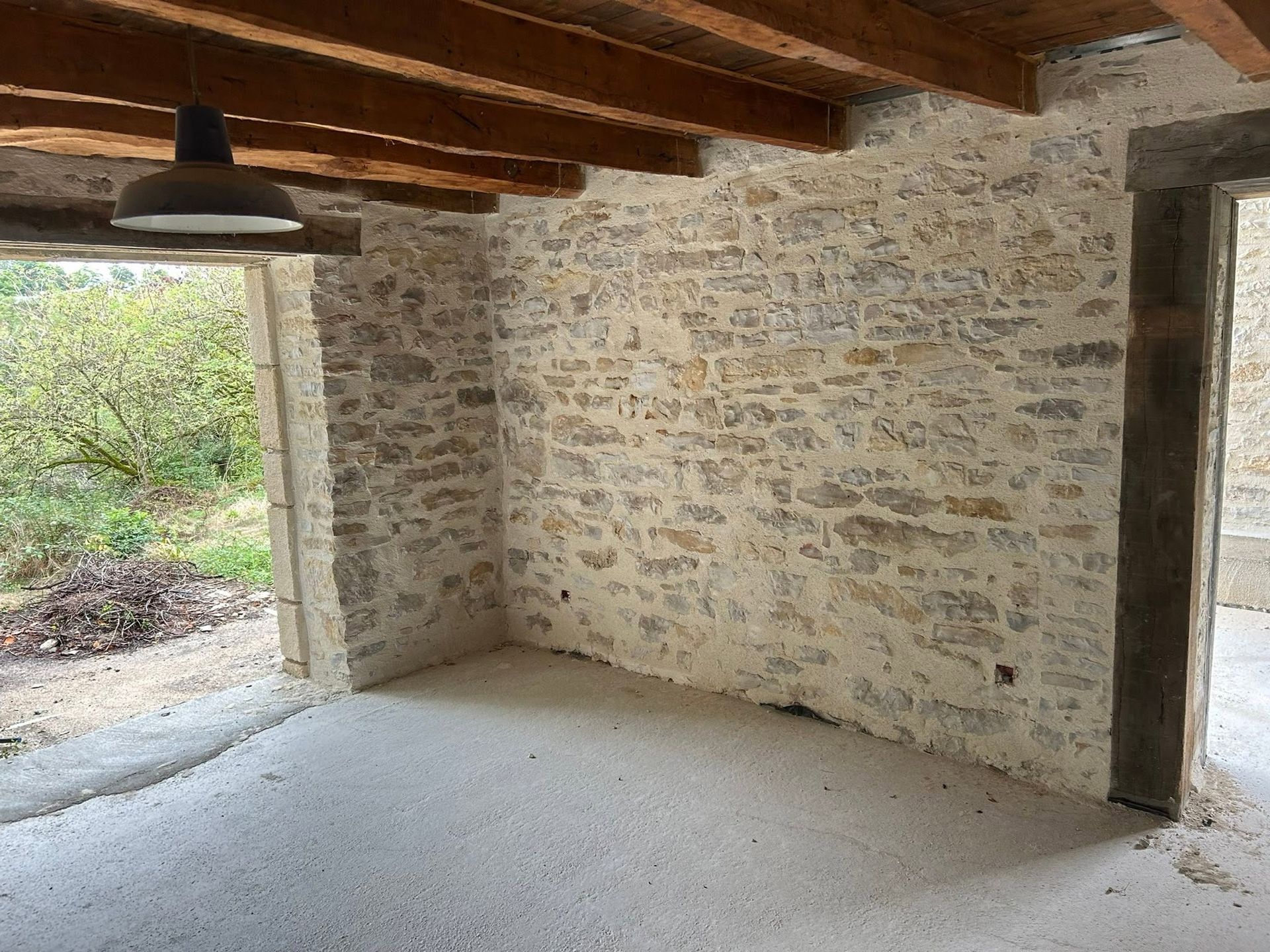 Pièce rustique aux murs en pierre, poutres apparentes et suspension lumineuse, s'ouvrant sur l'extérieur.