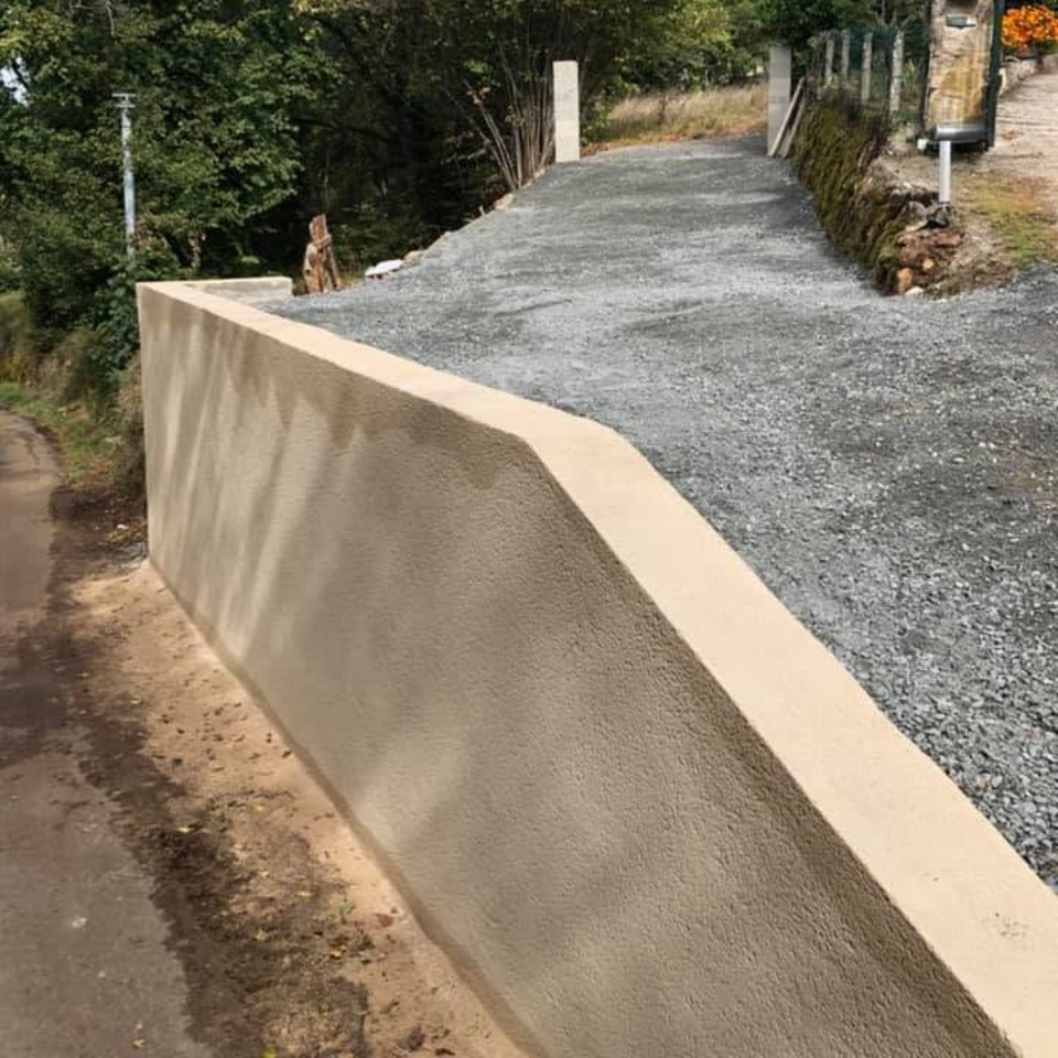 Mur de soutènement en béton bordant une allée de gravier sur un chemin en pente, avec des arbres