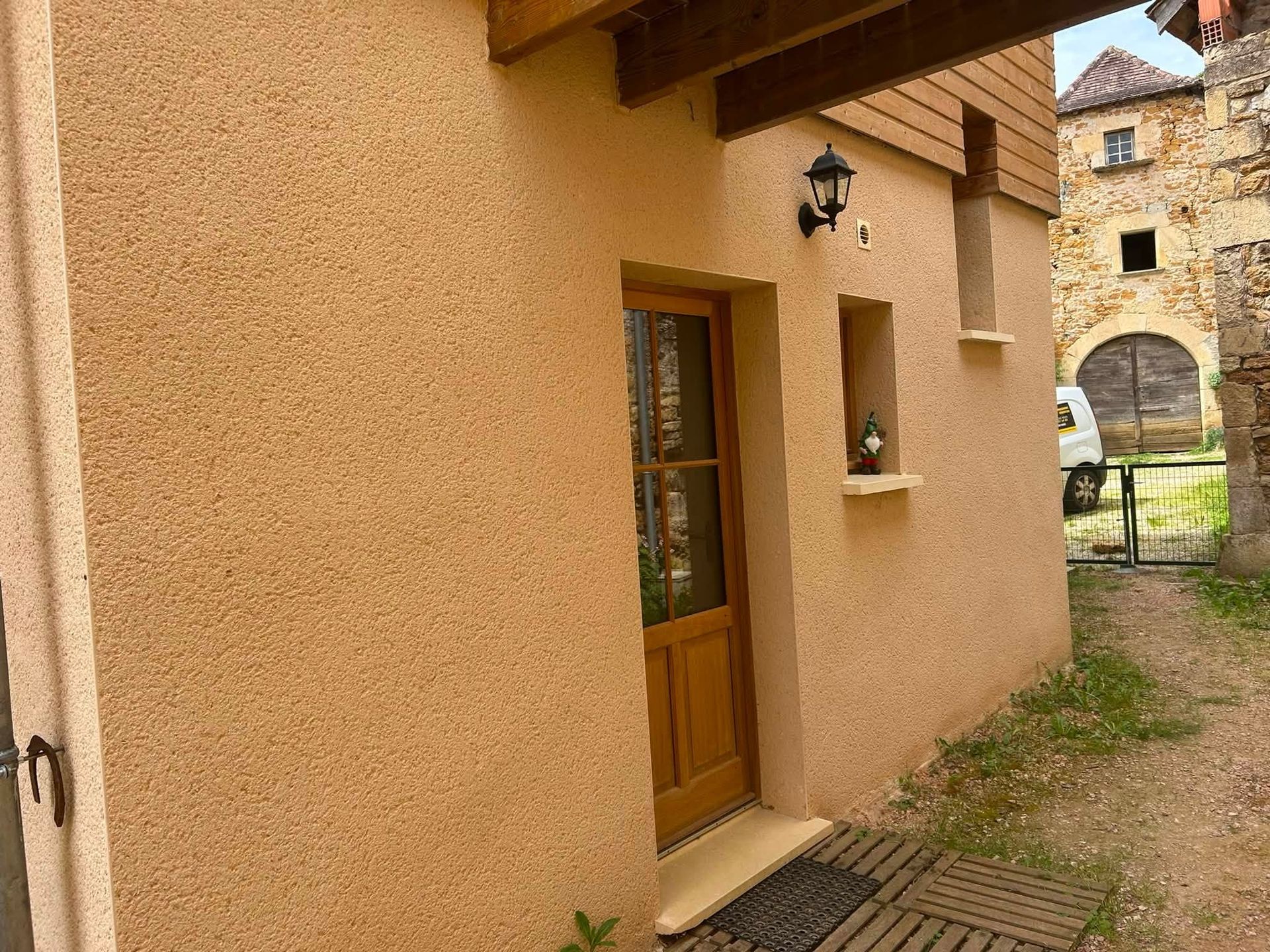 Façade d'un bâtiment beige avec porte en bois, applique murale et chemin vers une arche en pierre.