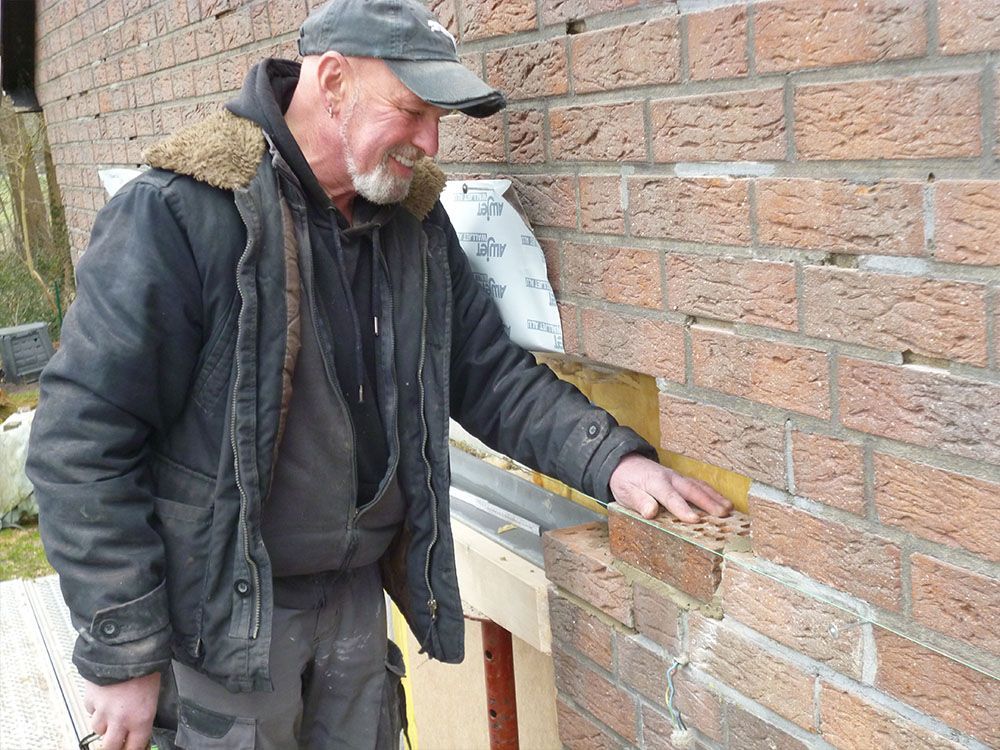 Ein Mann in einer schwarzen Jacke und einem schwarzen Hut steht neben einer Backsteinmauer.